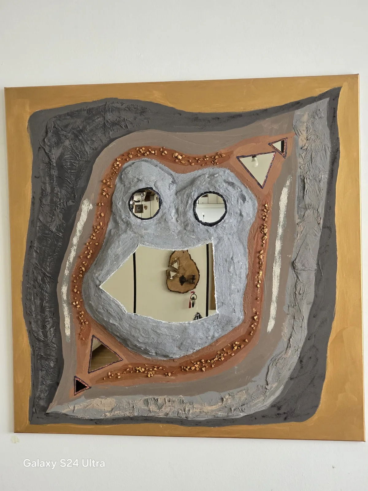 Reliefartiges Wandobjekt in Grau, Terrakotta und Gold mit eingelassenen Spiegelflächen, die als Augen und Dreiecke gesetzt sind.